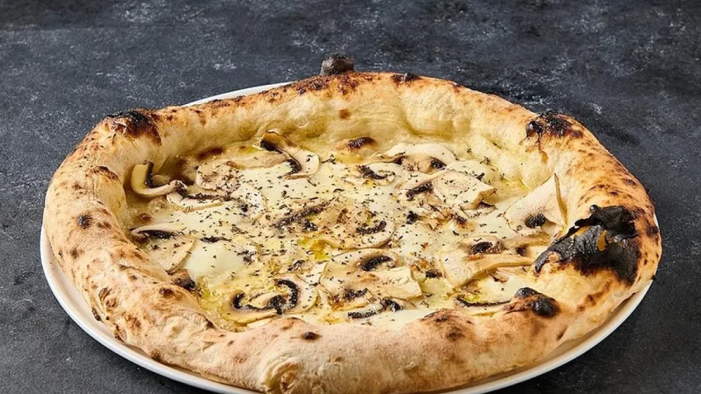 Pizza bianca com cogumelos laminados e azeite sobre prato branco em superfície de pedra escura.