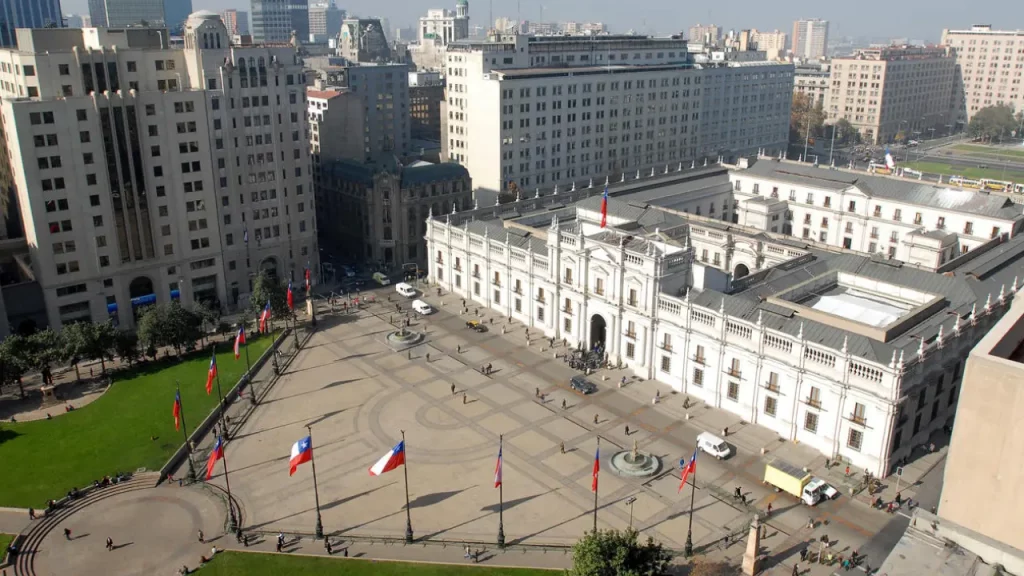 Vista aérea do Palácio de La Moneda no Centro Histórico de Santiago, com bandeiras chilenas e pessoas caminhando pela ampla praça.