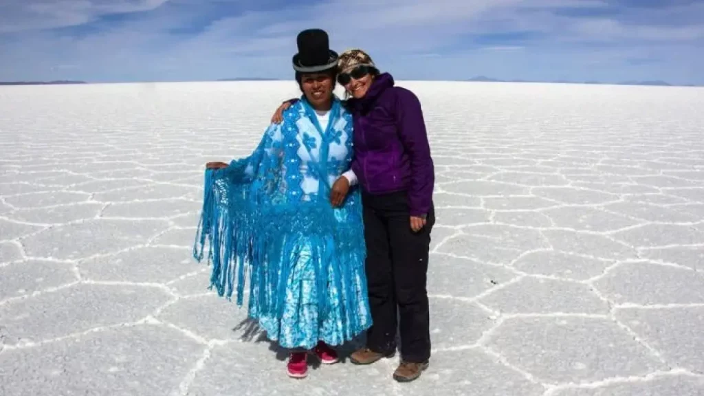 Duas mulheres abraçadas no Salar de Uyuni; uma veste traje andino tradicional azul com chapéu-coco, e a outra usa roupa de trilha. O chão branco do salar forma padrões hexagonais sob um céu azul.