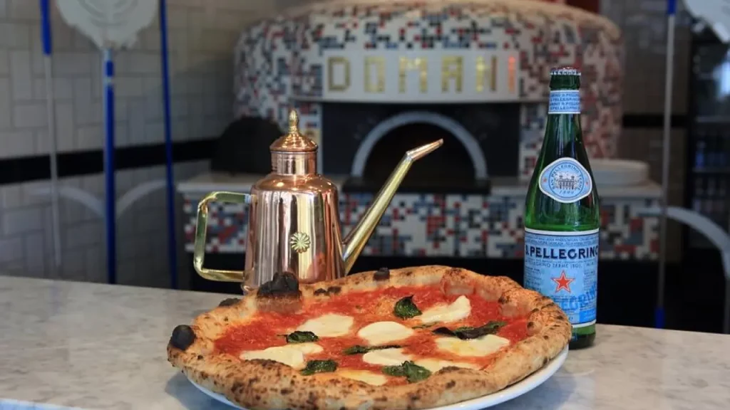 Pizza Margherita napolitana sobre balcão de mármore, forno mosaico “Domani” ao fundo, galheteiro de cobre e garrafa San Pellegrino ao lado.