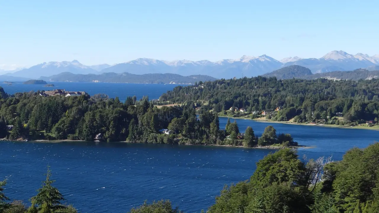 Vista panorâmica de um lago azul intenso rodeado por florestas e montanhas na região de Bariloche, com construções espalhadas entre as árvores e cordilheira ao fundo.