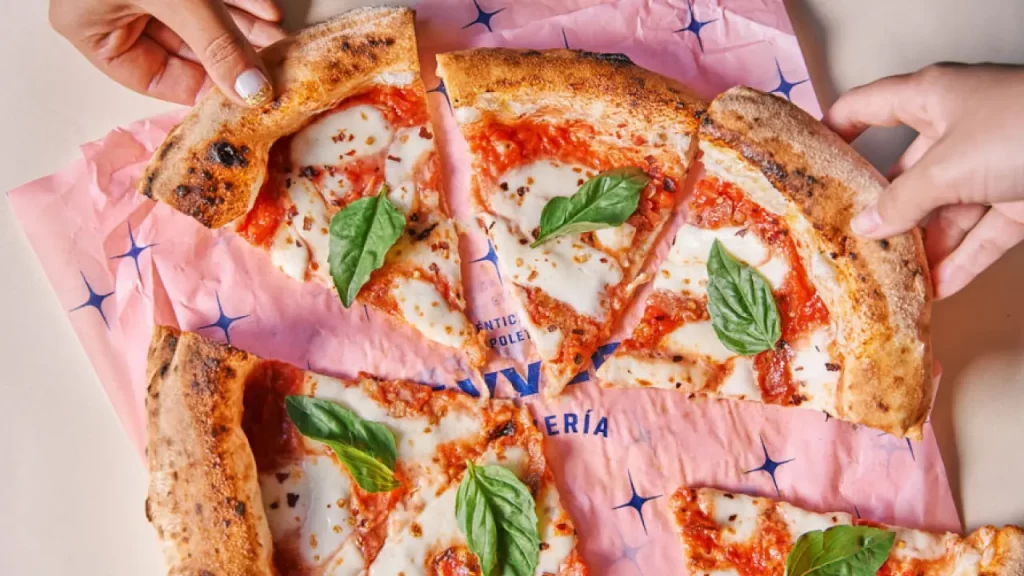 Pizza Margherita fatiada sobre papel rosa da pizzaria, queijo derretido e folhas frescas de manjericão.