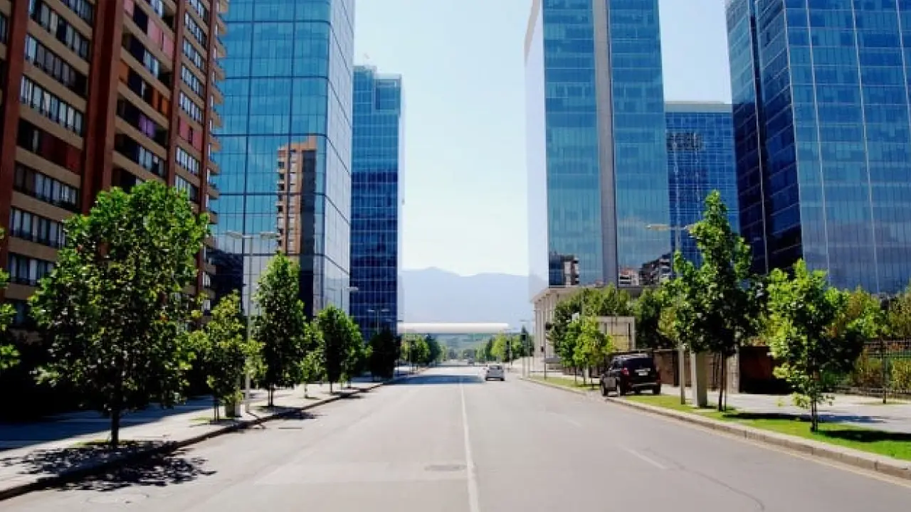 Avenida moderna e arborizada em Las Condes, Santiago, com prédios comerciais de vidro espelhado e vista para as montanhas ao fundo.