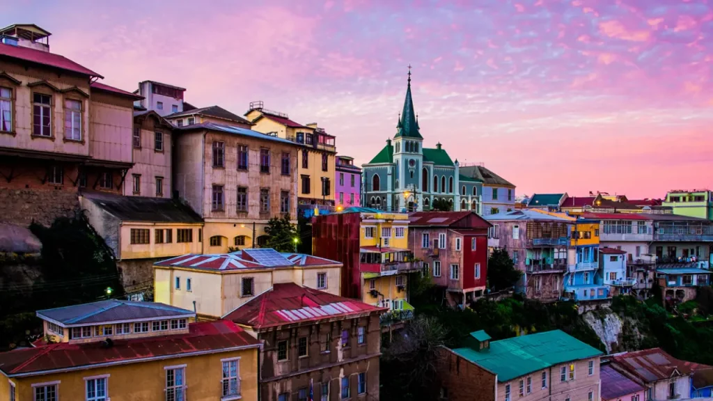 Casas coloridas nos morros de Valparaíso durante o pôr do sol, com céu rosa e igreja ao centro.