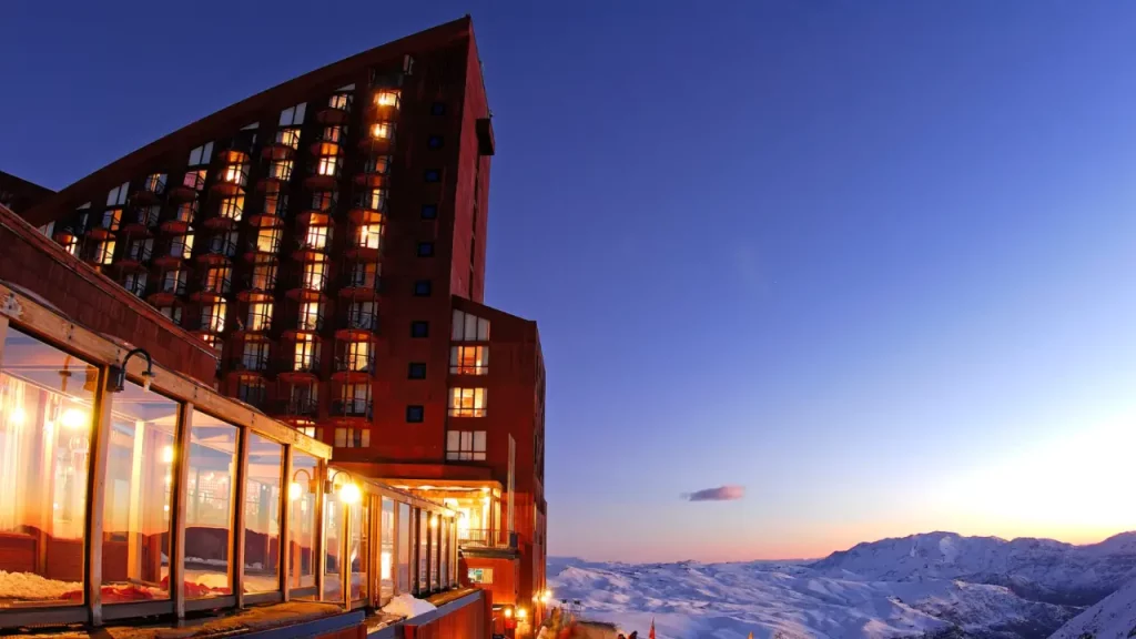 Hotel nas montanhas de Valle Nevado iluminado ao entardecer, com vista para a neve e turistas reunidos na entrada.