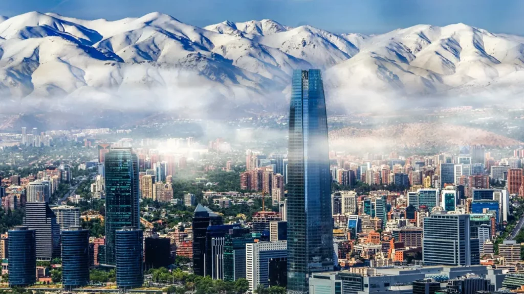 Vista panorâmica da cidade de Santiago, Chile, com o arranha-céu Costanera Center em destaque e a Cordilheira dos Andes coberta de neve ao fundo.