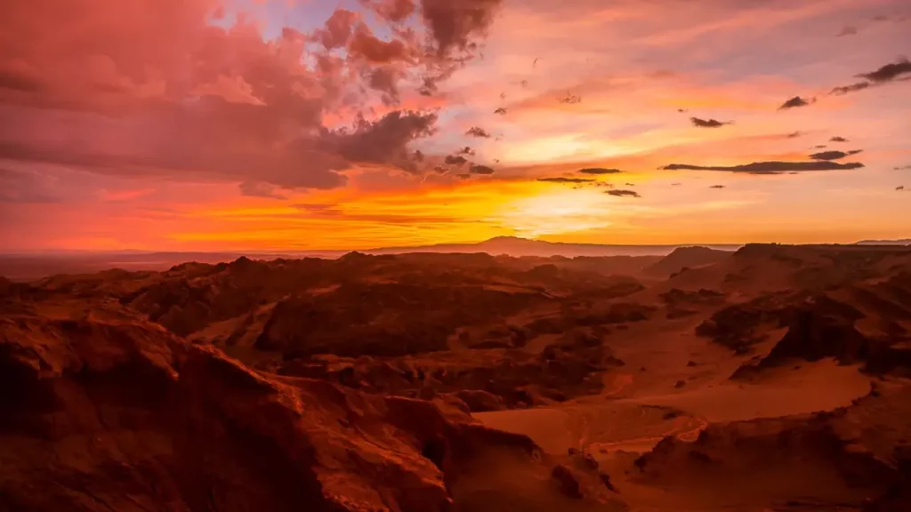 Fim de tarde no Valle de la Luna, no Deserto do Atacama, com céu alaranjado, formações rochosas e luz dourada refletindo na paisagem.