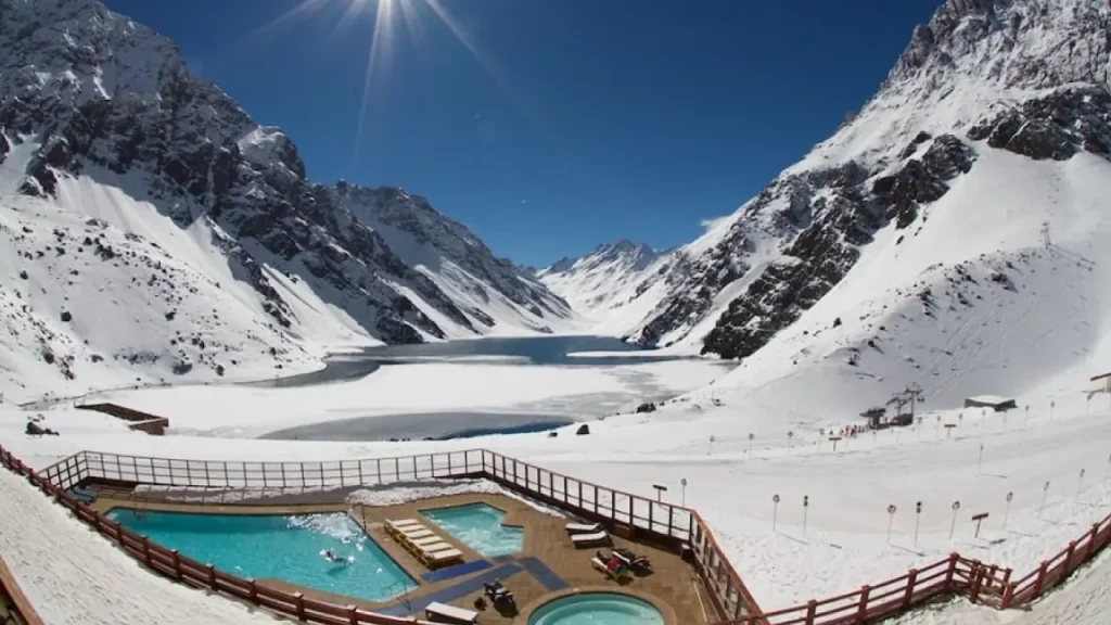 Vista panorâmica da estação de esqui Portillo no Chile com lago congelado, montanhas nevadas e piscinas externas sob o sol do inverno.