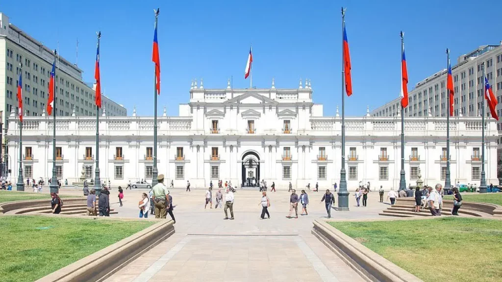 Fachada do Palácio de La Moneda em Santiago do Chile, com bandeiras chilenas ao redor e pessoas caminhando pela Plaza de la Ciudadanía em um dia ensolarado.