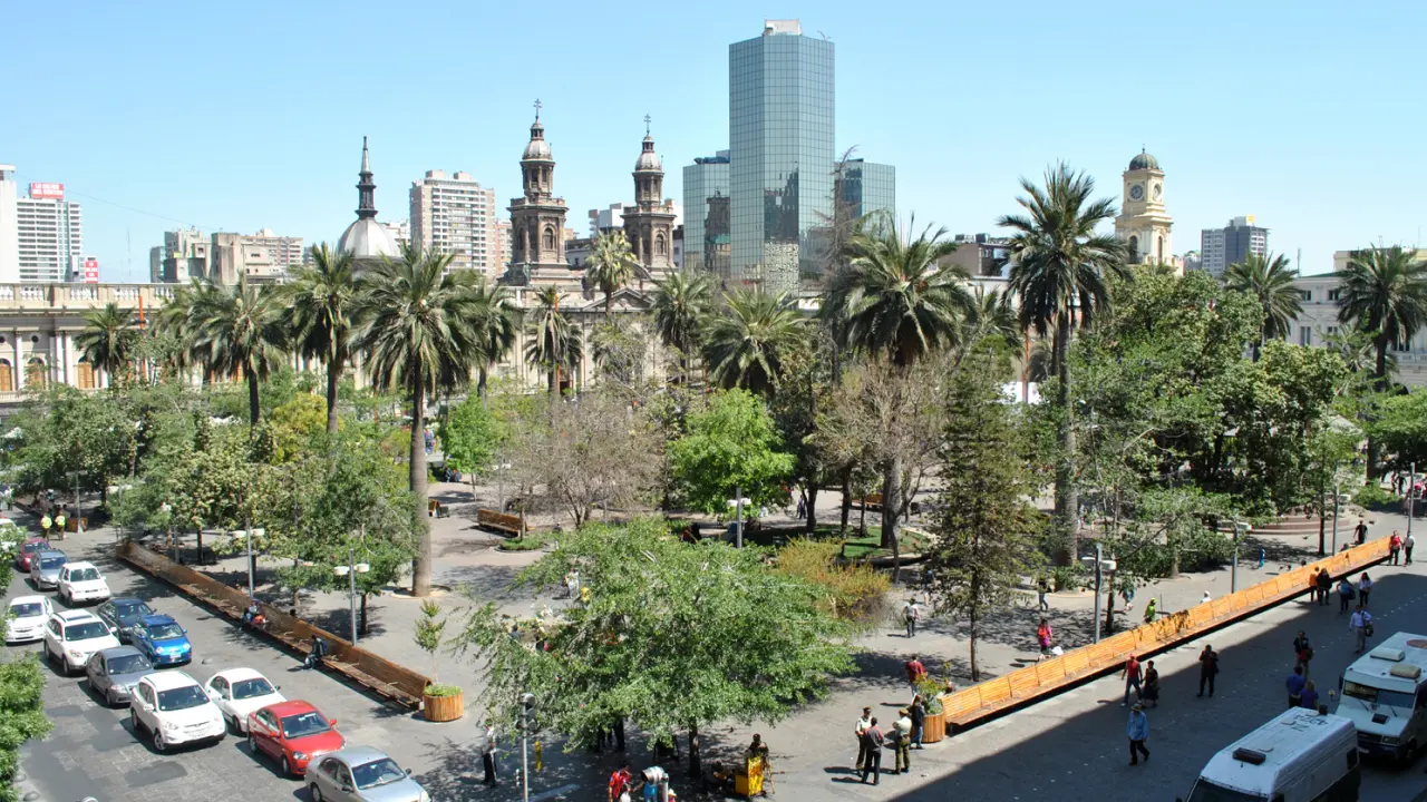 Vista panorâmica da Plaza de Armas em Santiago do Chile com palmeiras, prédios históricos e pessoas passeando.