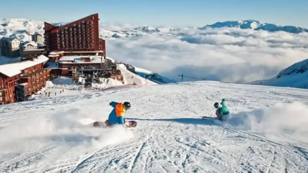 Dois snowboarders descem uma montanha nevada em alta velocidade, com o Hotel Valle Nevado e um mar de nuvens ao fundo.