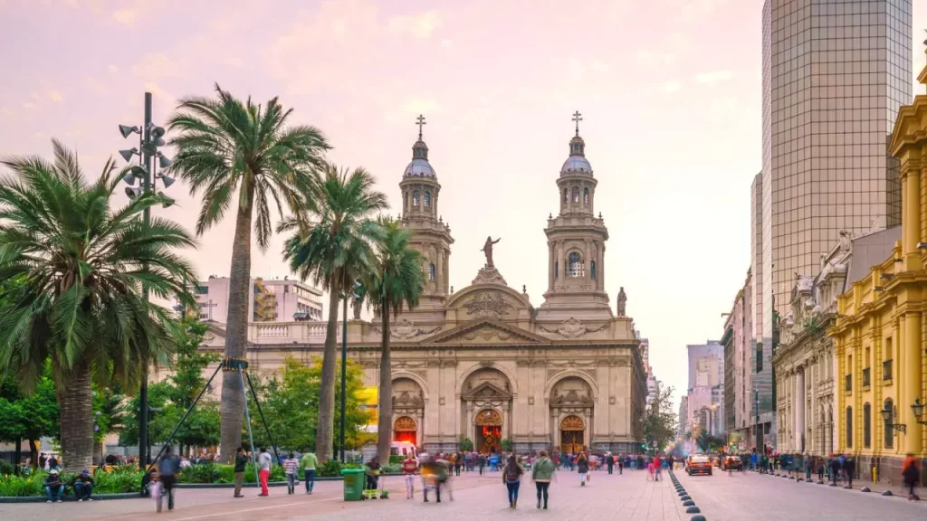 Vista da Catedral Metropolitana de Santiago ao entardecer, com palmeiras ao redor e movimento de pessoas na Plaza de Armas.