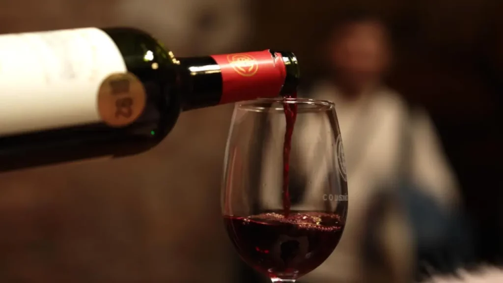 Vinho tinto sendo servido de uma garrafa para uma taça, em ambiente aconchegante com iluminação suave e fundo desfocado.