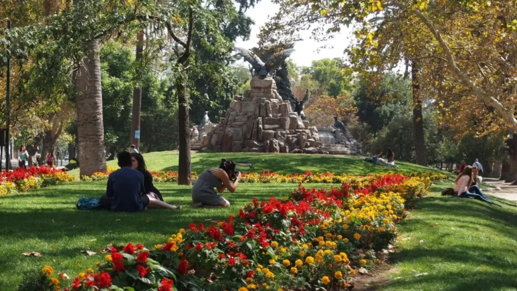 Parque arborizado com jardim florido e gramado verde. Pessoas estão sentadas ou deitadas na grama, aproveitando o ambiente natural e ensolarado. Ao fundo, uma grande escultura de pedra com figuras em bronze, incluindo uma águia com asas abertas, adorna o centro do parque.