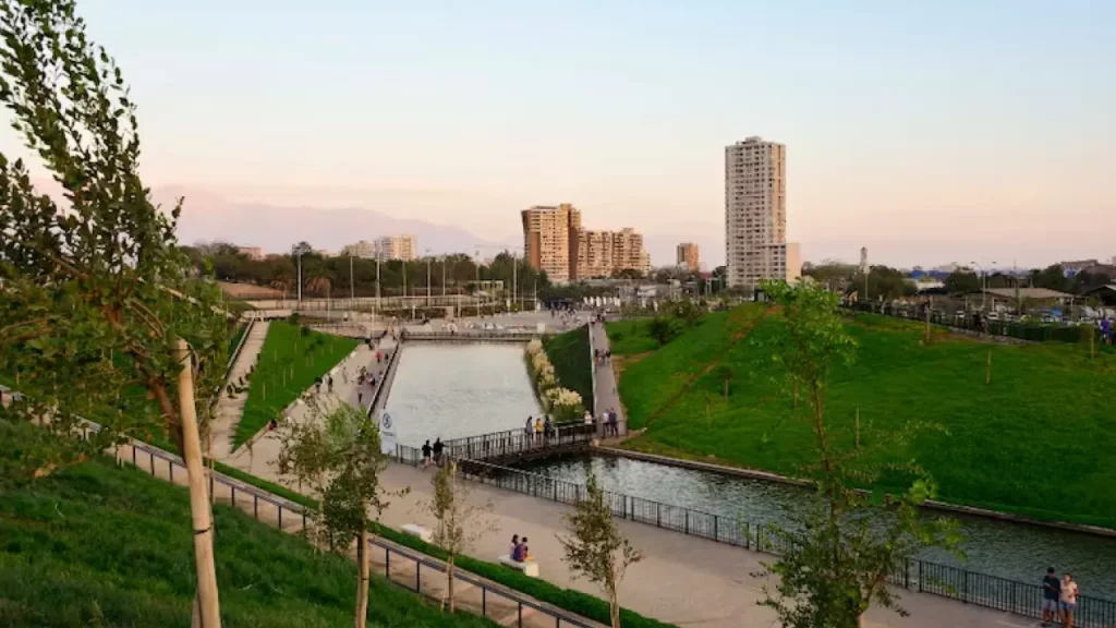 Parque urbano moderno com caminhos pavimentados e um canal central cercado por gramados bem cuidados. Diversas pessoas caminham e relaxam pelo local. Ao fundo, vê-se edifícios altos e uma leve névoa cobrindo as montanhas, sob um céu claro ao entardecer.
