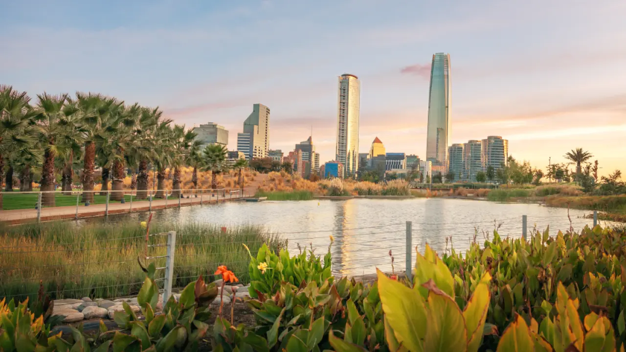 Vista de um parque com lago em primeiro plano, vegetação variada e palmeiras alinhadas. Ao fundo, destaca-se o skyline moderno da cidade, com prédios altos e o imponente edifício Costanera Center, refletido na água. O céu exibe tons suaves de pôr do sol.