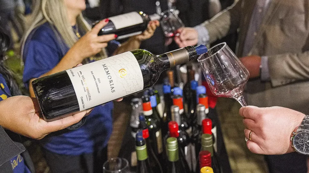 Pessoa servindo vinho tinto da garrafa Memorias em uma taça durante evento de degustação, com várias garrafas ao fundo.