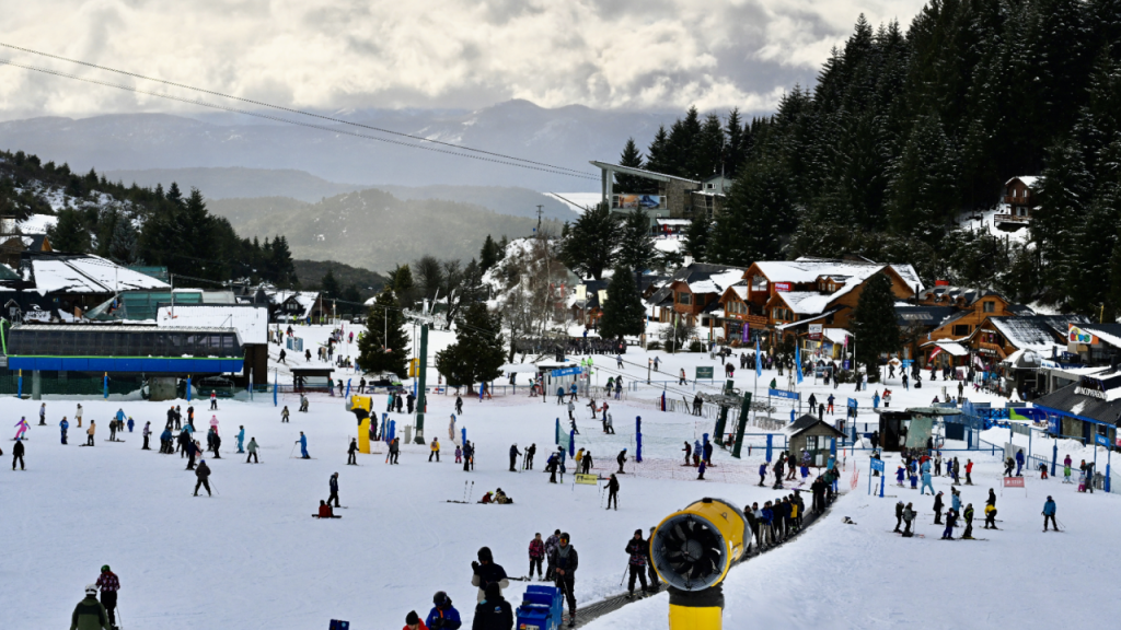 Panorâmica da área central do Cerro Catedral em Bariloche, com visitantes, teleféricos e construções típicas cercadas por floresta de pinheiros.