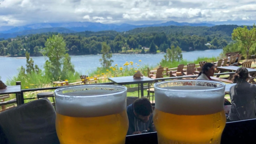 Duas taças de cerveja vistas de uma mesa com ambiente ao ar livre, em frente ao lago Nahuel Huapi, rodeado por vegetação e montanhas, com outras mesas ocupadas ao fundo.