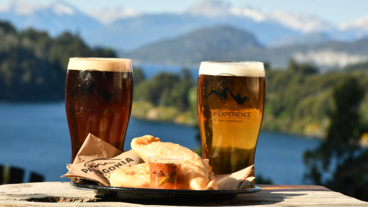 Duas taças de cerveja artesanal com logotipo da Bariloche Beer Experience ao ar livre, acompanhadas de empanadas e molho, com vista para o lago e montanhas ao fundo.