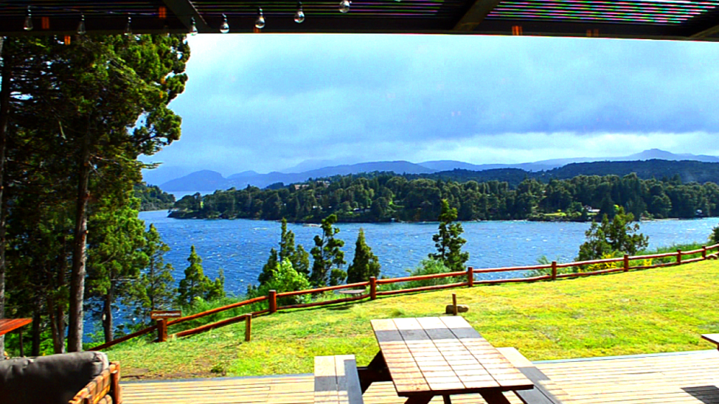 Área externa com bancos de madeira vazios voltados para o lago e montanhas de Bariloche, cercada por árvores e gramado verde, sob um céu nublado.
