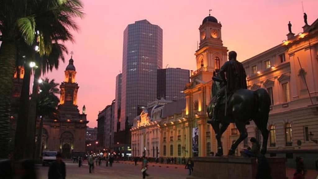 Praça de Armas de Santiago ao pôr do sol, com a Catedral Metropolitana iluminada e edifícios históricos no centro da cidade.