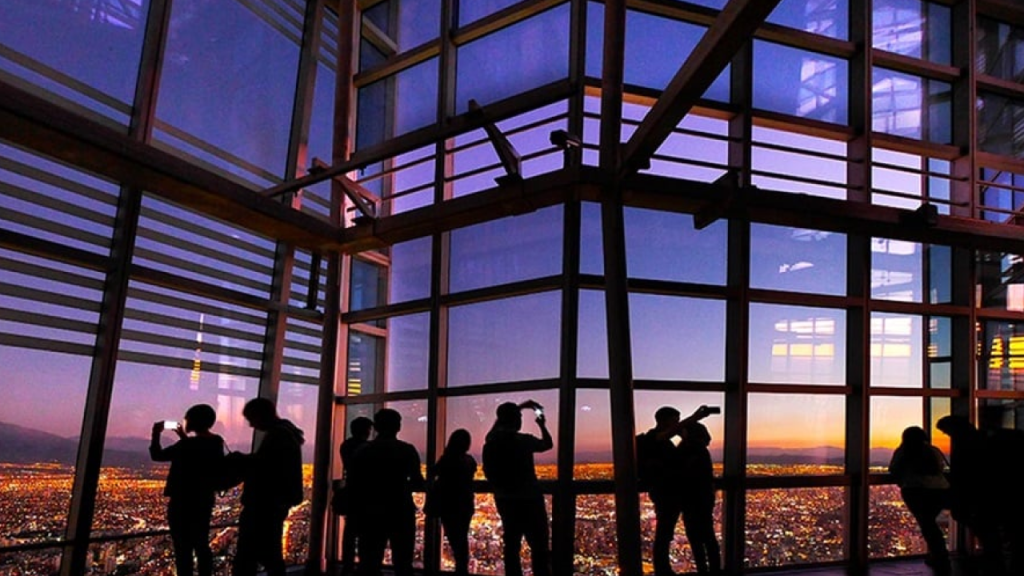 Visitantes observando o pôr do sol no mirante Sky Costanera, com janelas panorâmicas e vista da cidade de Santiago iluminada.