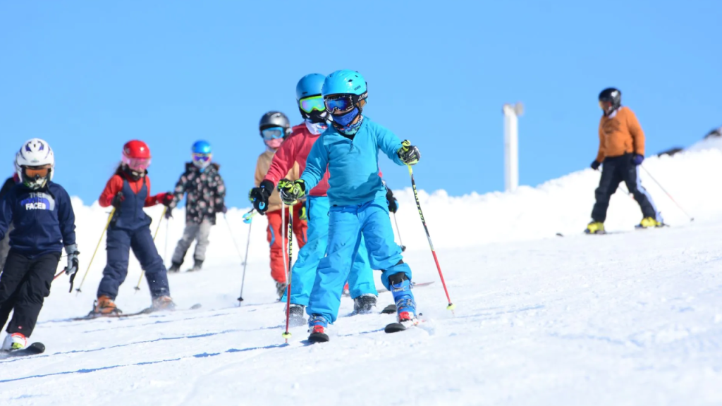Grupo de crianças aprendendo a esquiar em Bariloche, em uma pista de neve sob céu azul, usando roupas e equipamentos de esqui coloridos.