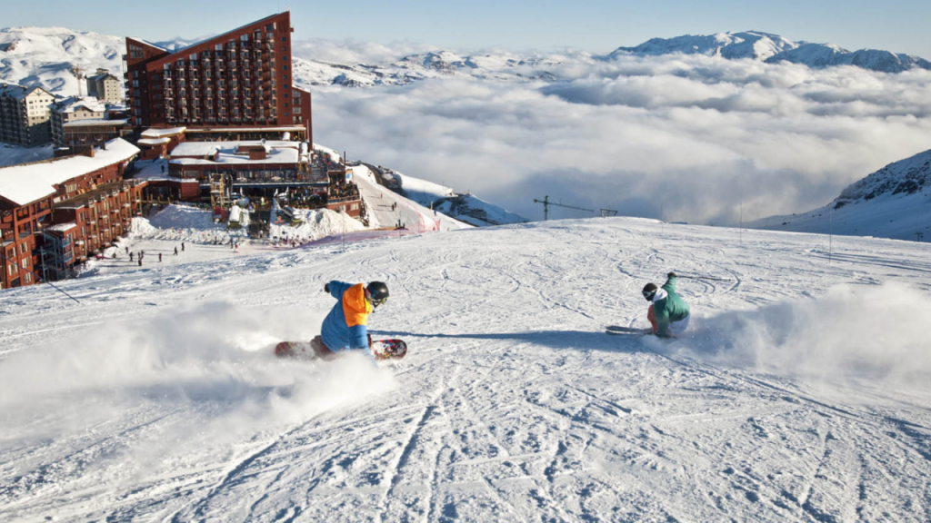 Crianças praticando snowboard no Valle Nevado, com vista panorâmica das montanhas nevadas e do complexo hoteleiro ao fundo.