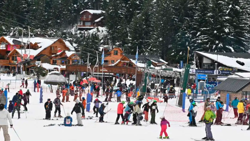 Crianças e adultos se divertindo na base da estação de esqui Cerro Catedral em Bariloche, com prédios de madeira ao fundo e neve por todo lado.