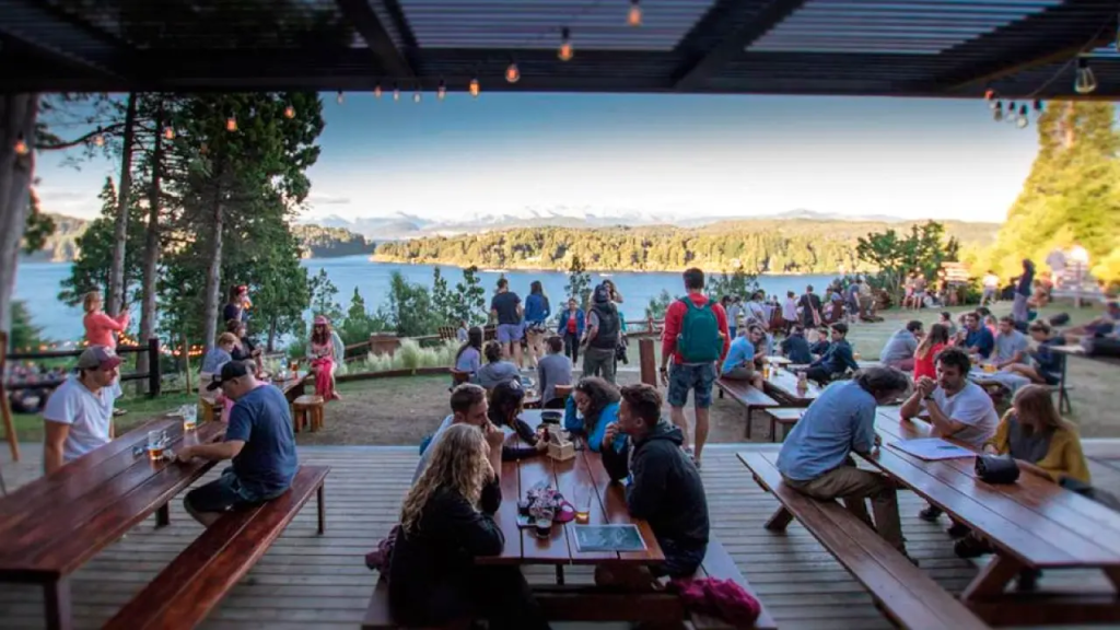 Pessoas reunidas em mesas de madeira no tour cervejeiro de Bariloche, aproveitando a vista para o lago e as montanhas ao entardecer, em ambiente descontraído ao ar livre.