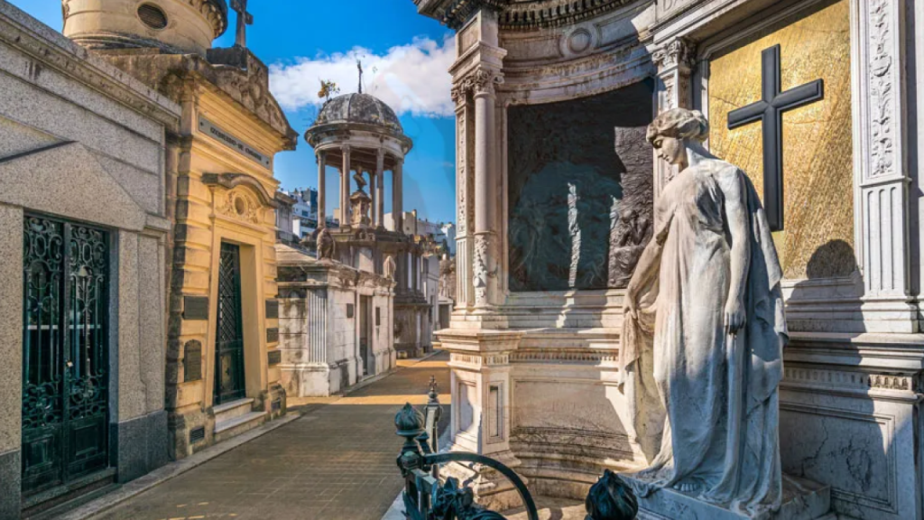 Corredor com mausoléus ornamentados e esculturas no Cemitério da Recoleta, céu azul ao fundo.