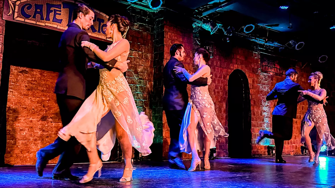 Casais dançando tango em palco de casa de shows portenha, figurinos elegantes e luz de espetáculo.