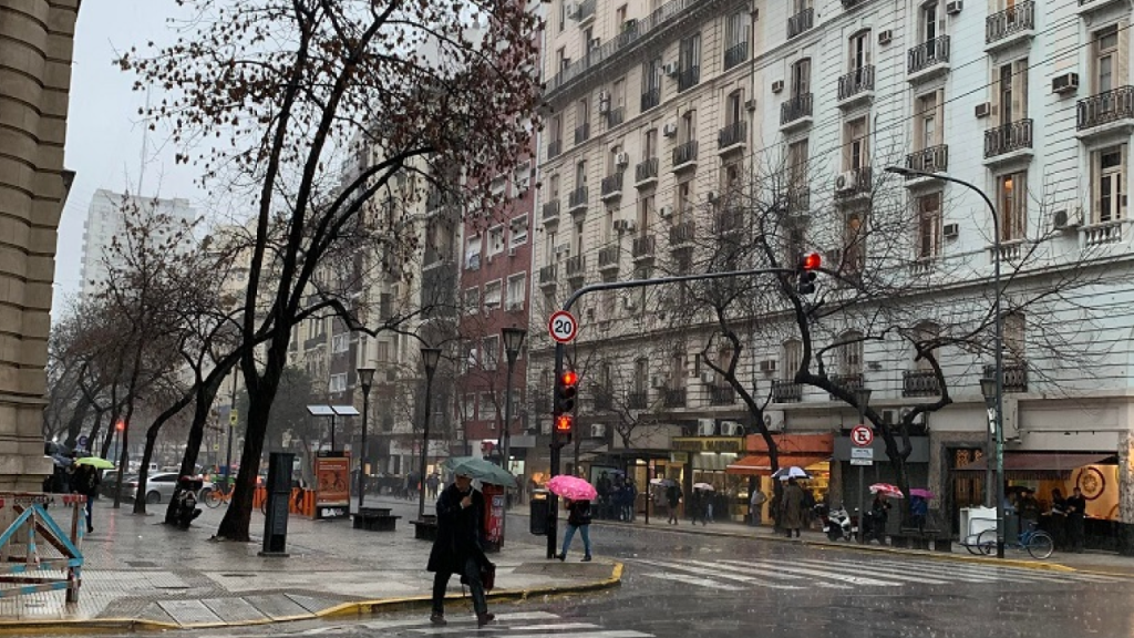 Rua de Buenos Aires no inverno com chuva, pedestres de guarda-chuva e árvores sem folhas.